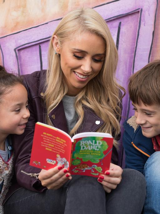 Eine blonde Frau liest zwei Kindern aus einem Buch von Roald Dahl vor.