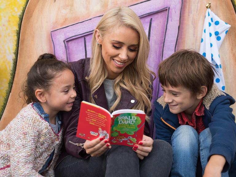 Eine blonde Frau liest zwei Kindern aus einem Buch von Roald Dahl vor.