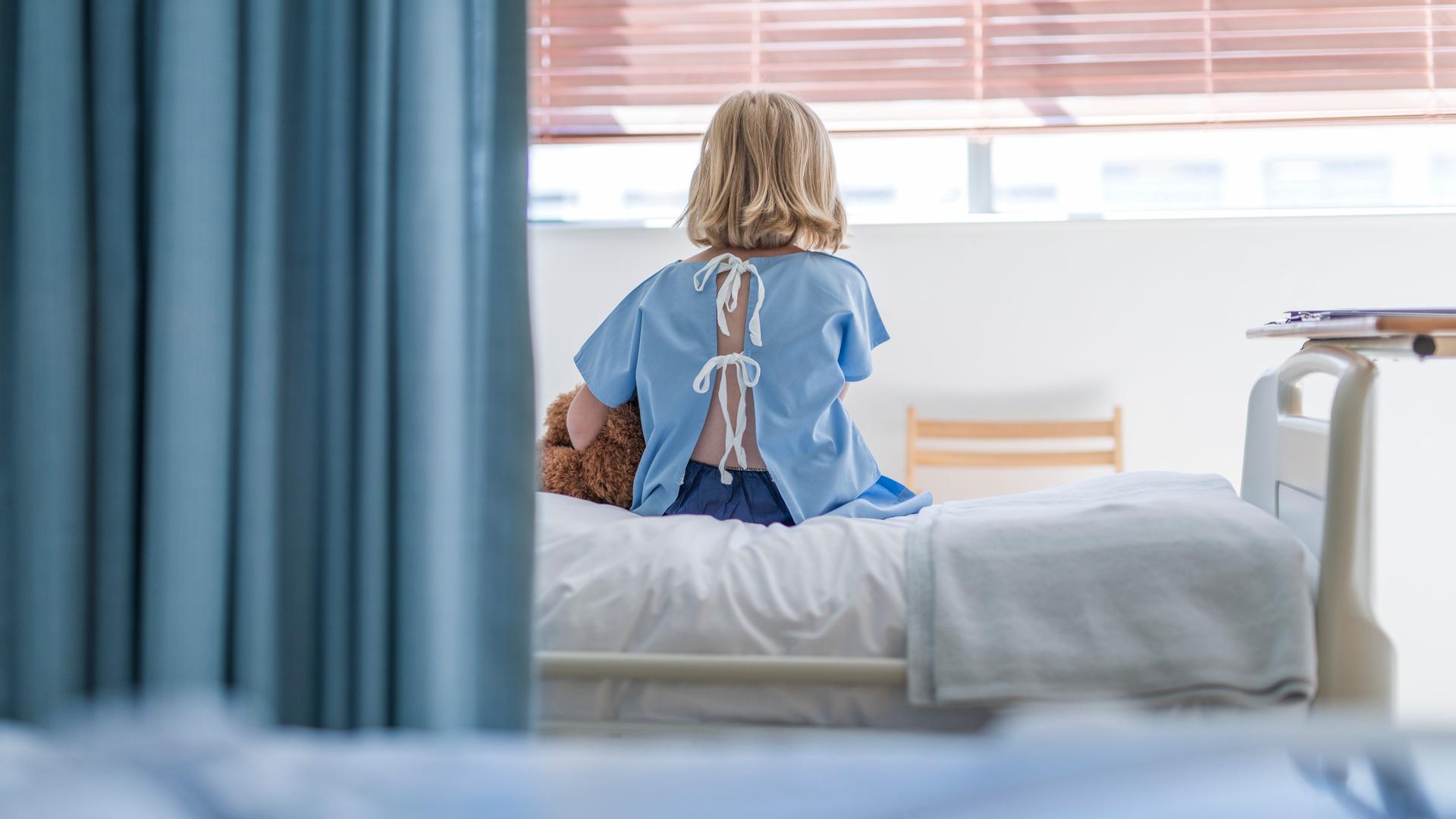 Rückansicht auf ein Kind, was auf einem Krankenhausbett sitzt, einen Teddy unter dem Arm.