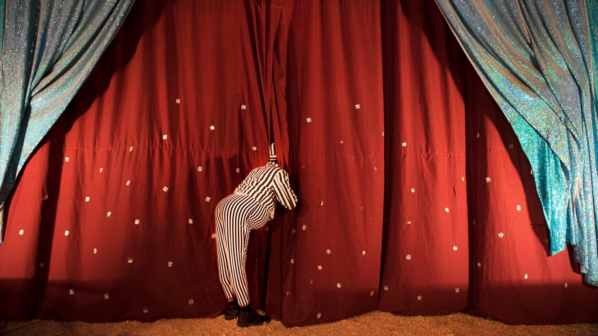 Ein Clown schaut hinter die Zirkusvorhänge, sodass sein Gesicht nicht zu sehen ist. Ein Clown schaut hinter die Zirkusvorhänge, sodass sein Gesicht nicht zu sehen ist.