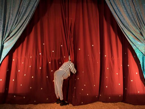 Ein Clown schaut hinter die Zirkusvorhänge, sodass sein Gesicht nicht zu sehen ist.