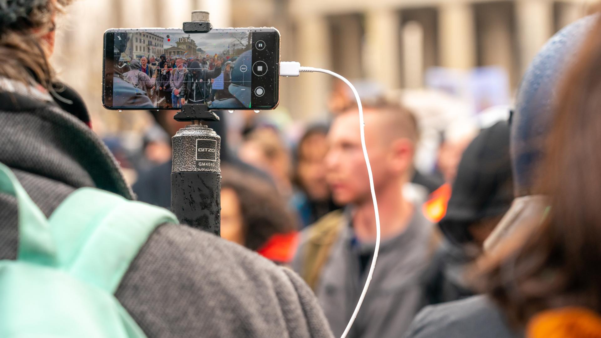 Ein Journalist steht mit einem Handy auf einem Stativ vor einer Demonstration gegen die Corona-Maßnahmen. 