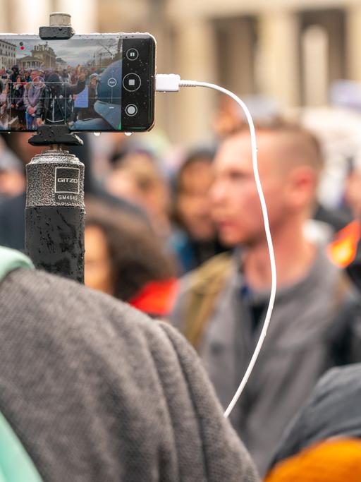 Ein Journalist steht mit einem Handy auf einem Stativ vor einer Demonstration gegen die Corona-Maßnahmen. 