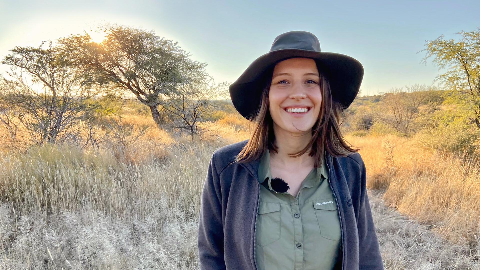 Eine junge Frau mit einem Hut steht, mit dem Rücken zur Sonne, mitten in einer Savannenlandschaften und lacht freundlich in die Kamera.