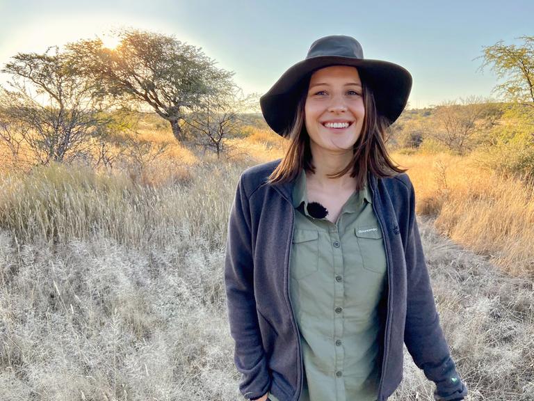 Eine junge Frau mit einem Hut steht, mit dem Rücken zur Sonne, mitten in einer Savannenlandschaften und lacht freundlich in die Kamera.