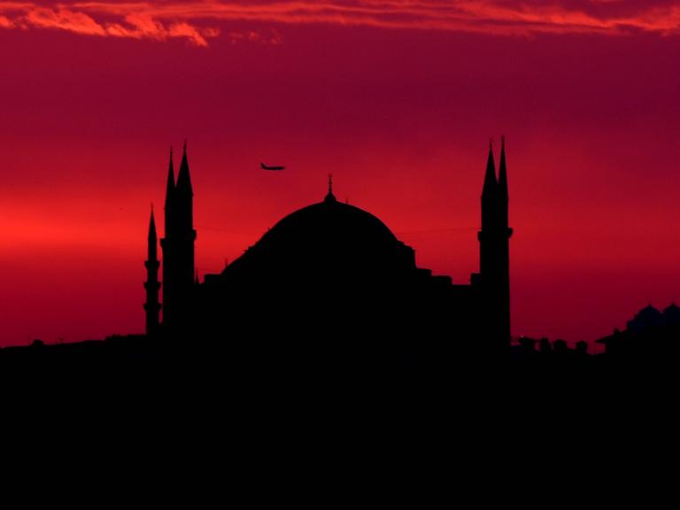 Die Shilouette der Hagia Sophia in Istanbul vor einem blutroten Himmel, an dem ein Flugzeug zu sehen ist.