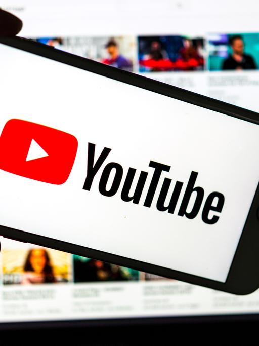 Das Logo des Video-Portals YouTube wird auf dem Display eines Smartphones angezeigt. Im Hintergrund ist auf einem Bildschirm die YouTube Homepage zu sehen.