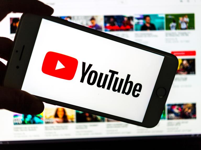 Das Logo des Video-Portals YouTube wird auf dem Display eines Smartphones angezeigt. Im Hintergrund ist auf einem Bildschirm die YouTube Homepage zu sehen.