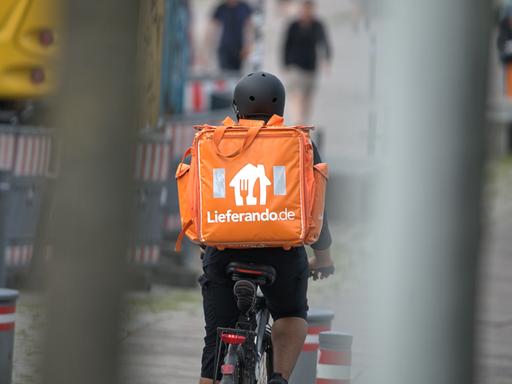 Ein Fahrradkurier des Fastfood-Lieferdienstes Lieferando.de mit Helm im Einsatz. Mit einem großen, orangefarbenen Thermo-Rucksack werden die bestellten Speisen per Fahrrad ausgeliefert.