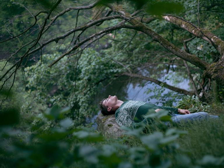 Ein Mann liegt mit geschlossenen Augen im Wald und meditiert.