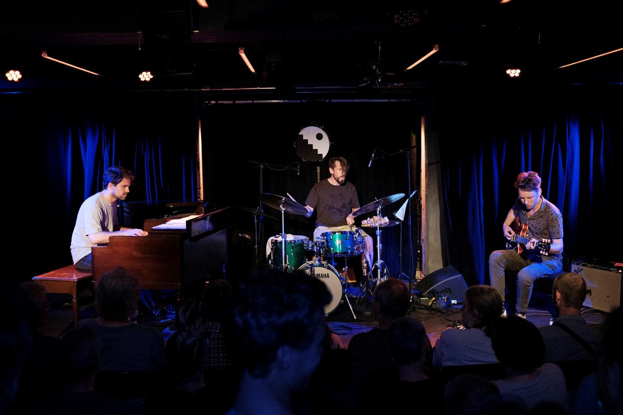 Drei Musiker sitzen auf einer kleinen Clubbühne mit Orgel, Schlagwerk und Gitarre und spielen gemeinsam. Drei Musiker sitzen auf einer kleinen Clubbühne mit Orgel, Schlagwerk und Gitarre und spielen gemeinsam.