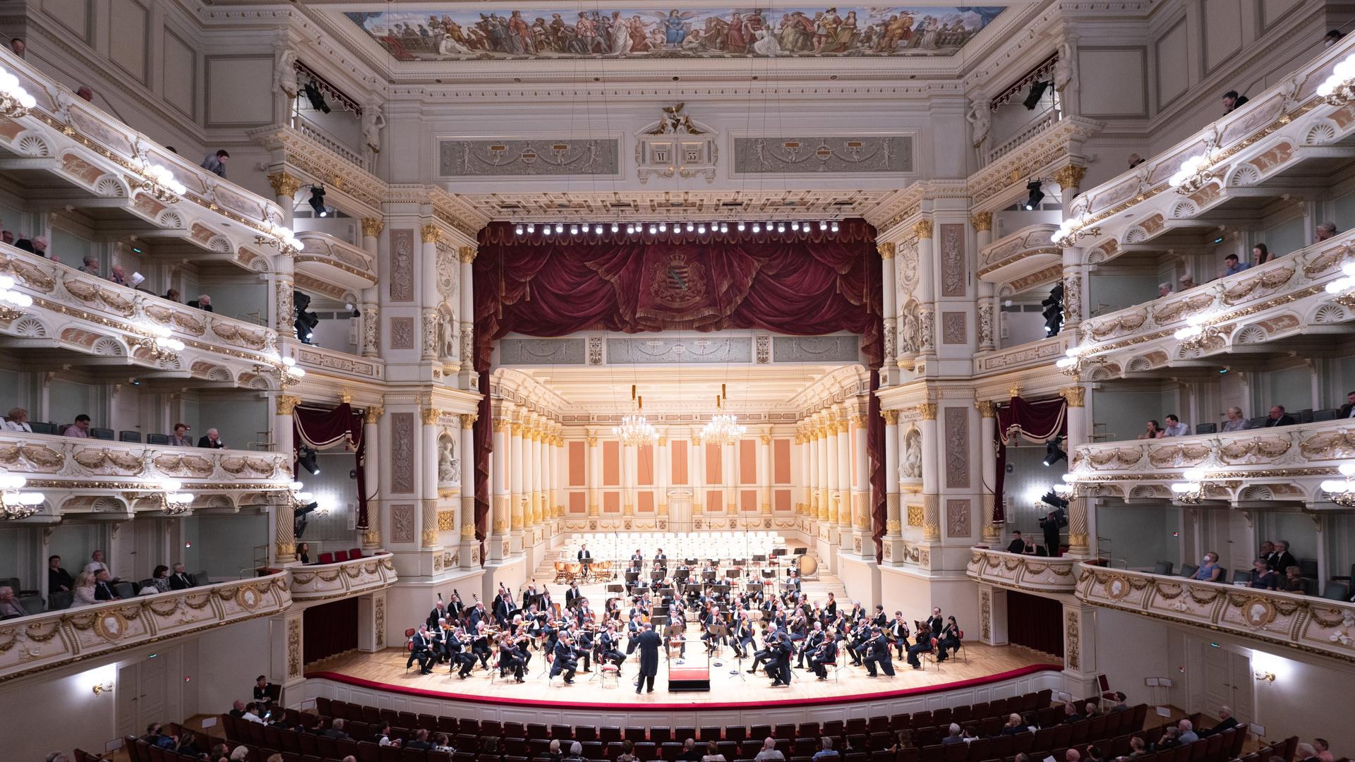 Ein Orchester sitzt in einem reichverzierten Konzertsaal.
