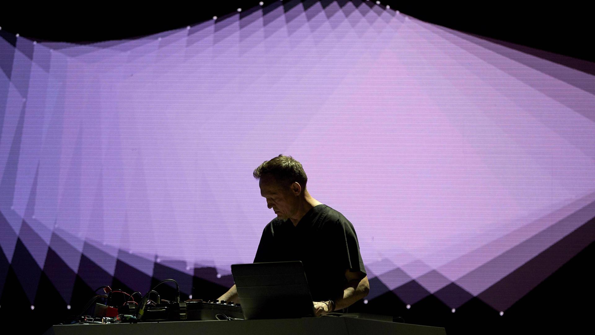 Ein Mann steht auf einer Bühne vor einem Laptop, im Hintergrund werden überlappende geometrische Formen in Lila projiziert.