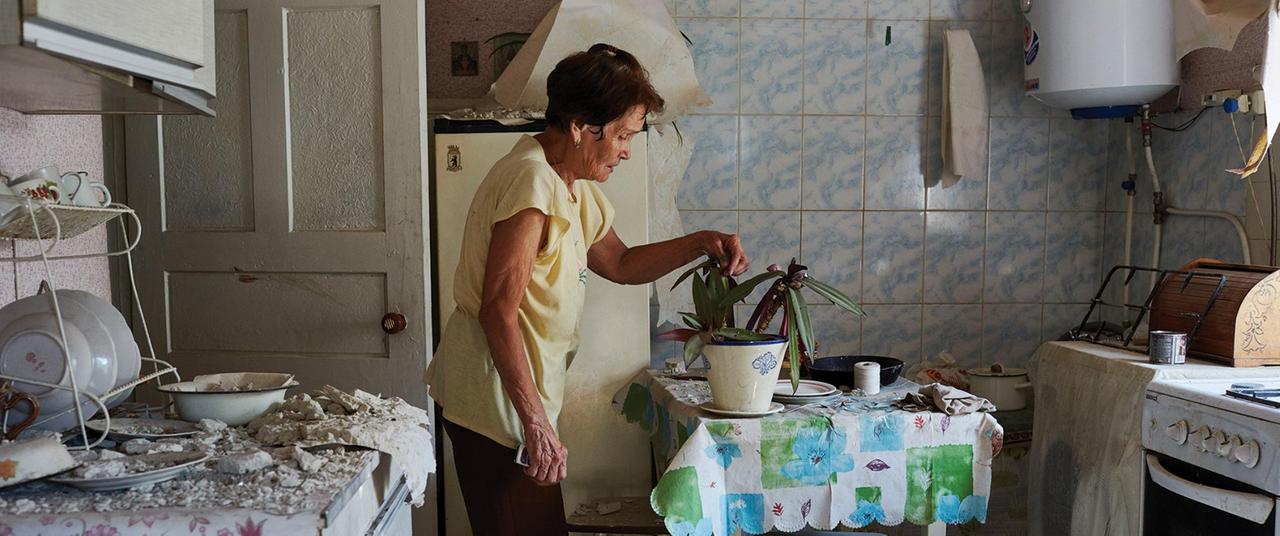Seitenansicht einer Frau, die sich in einer kargen Küche um eine Pflanze auf dem Esstisch kümmert. Auf dem Herd neben ihr sind herabgefallene Trümmer zu sehen.