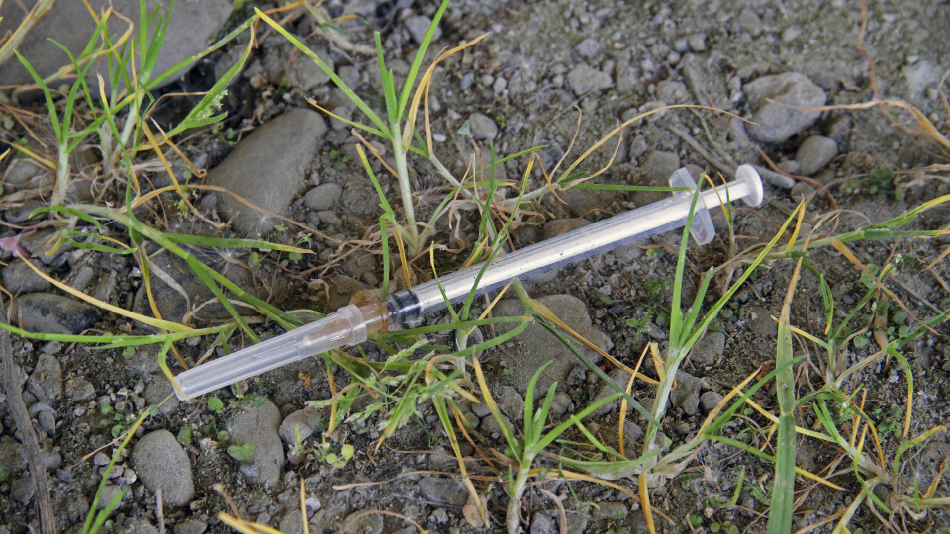Eine Heroinspritze liegt auf sandigem Untergrund, umgeben von Grasbüscheln. Eine Heroinspritze liegt auf sandigem Untergrund, umgeben von Grasbüscheln.