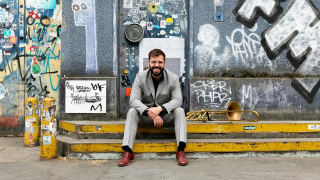Ein lachender Mann mit Brille und Vollbart sitzt auf den Stufen vor einer mit Graffiti bemalten Hauswand. Hinter ihm die mit Aufklebern übersäte Tür, neben ihm eine Posaune.