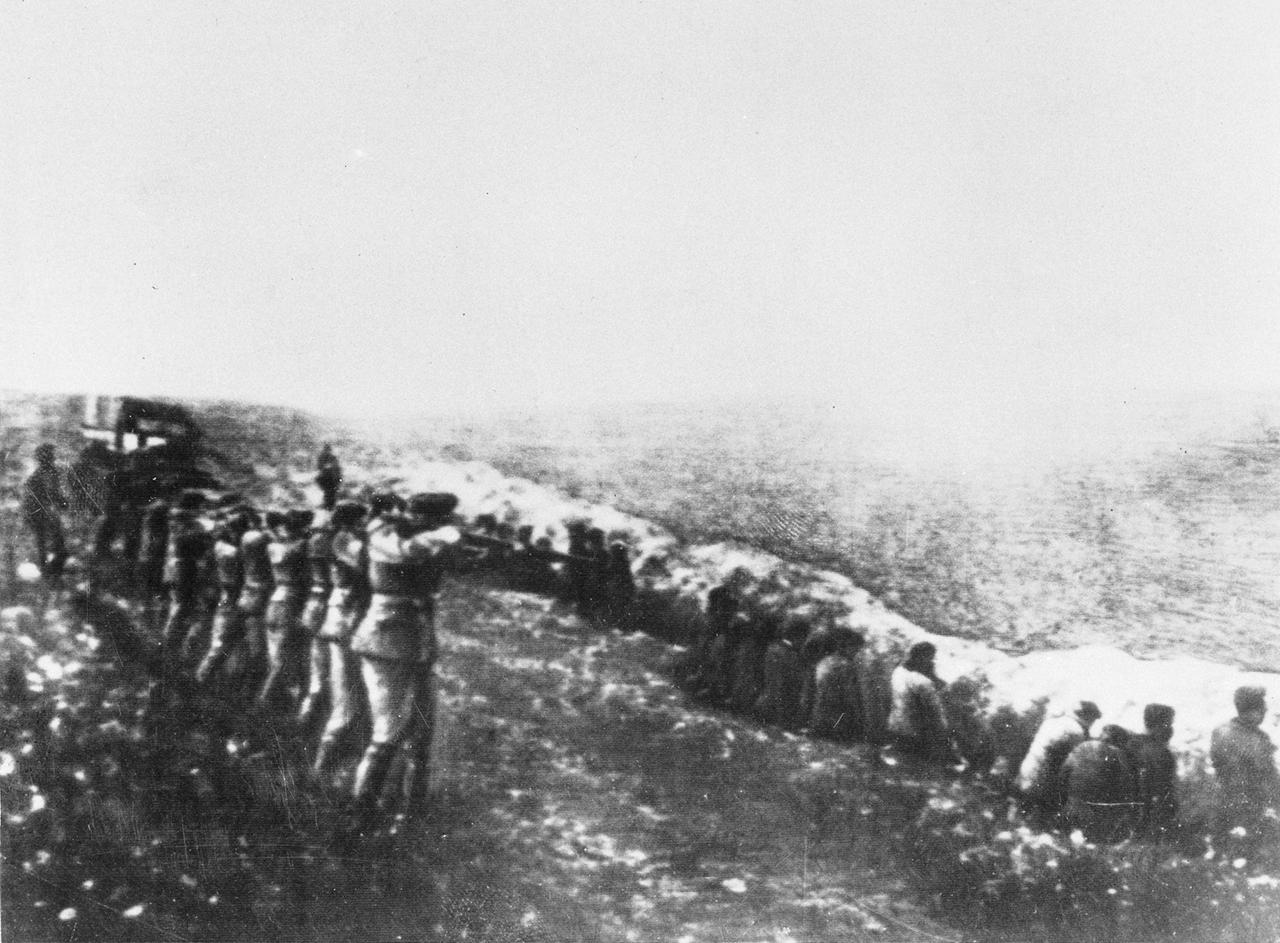 Historisches Foto von Wehrmachtssoldaten, die auf Zivilisten schießen, die mit dem Rücken zu den Soldaten in einer langen Reihe am Rande ihres eigenen Massengrabes sitzen.