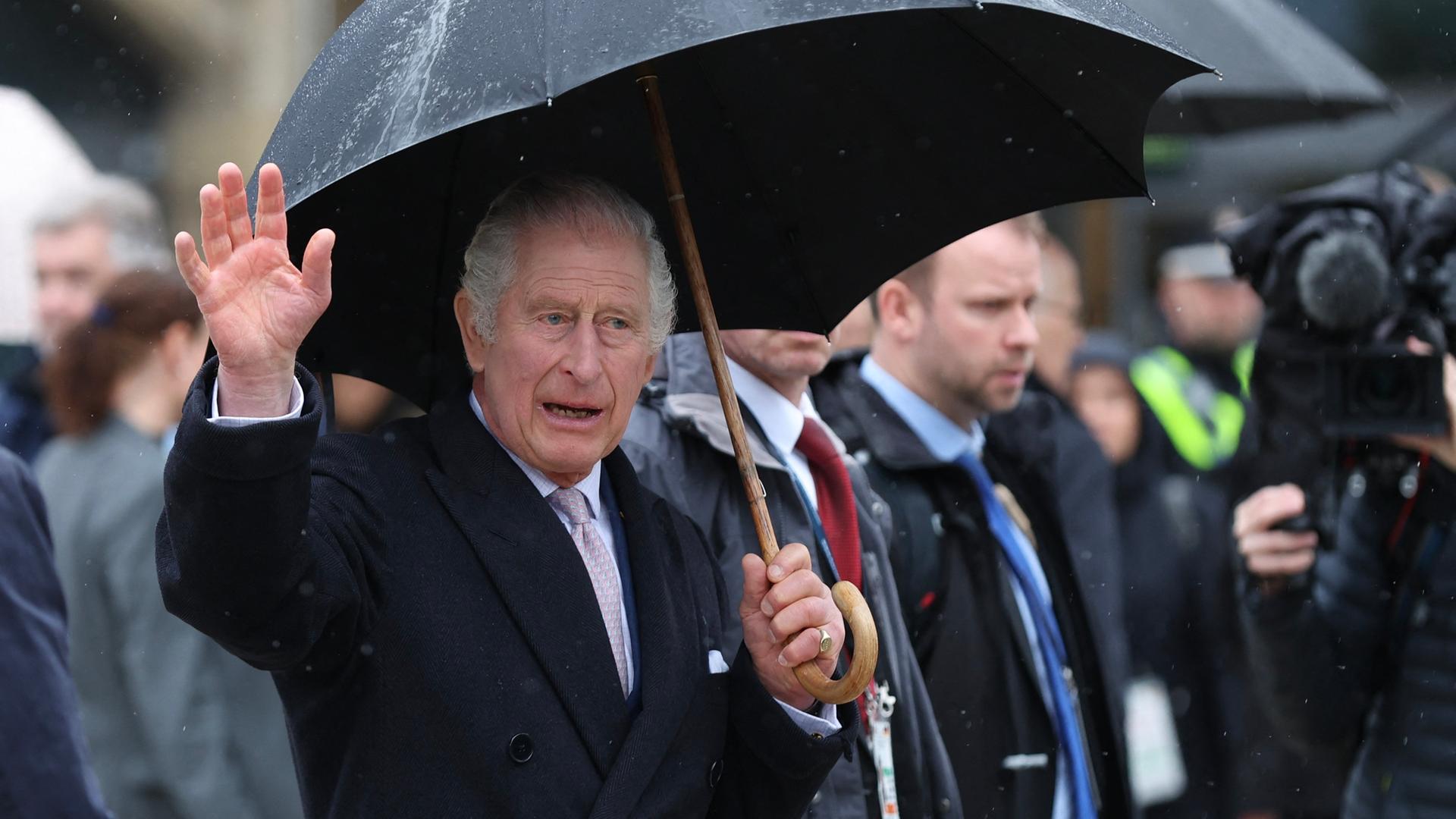 König Charles III. trägt einen Regenschirm und winkt den Menschen zu.