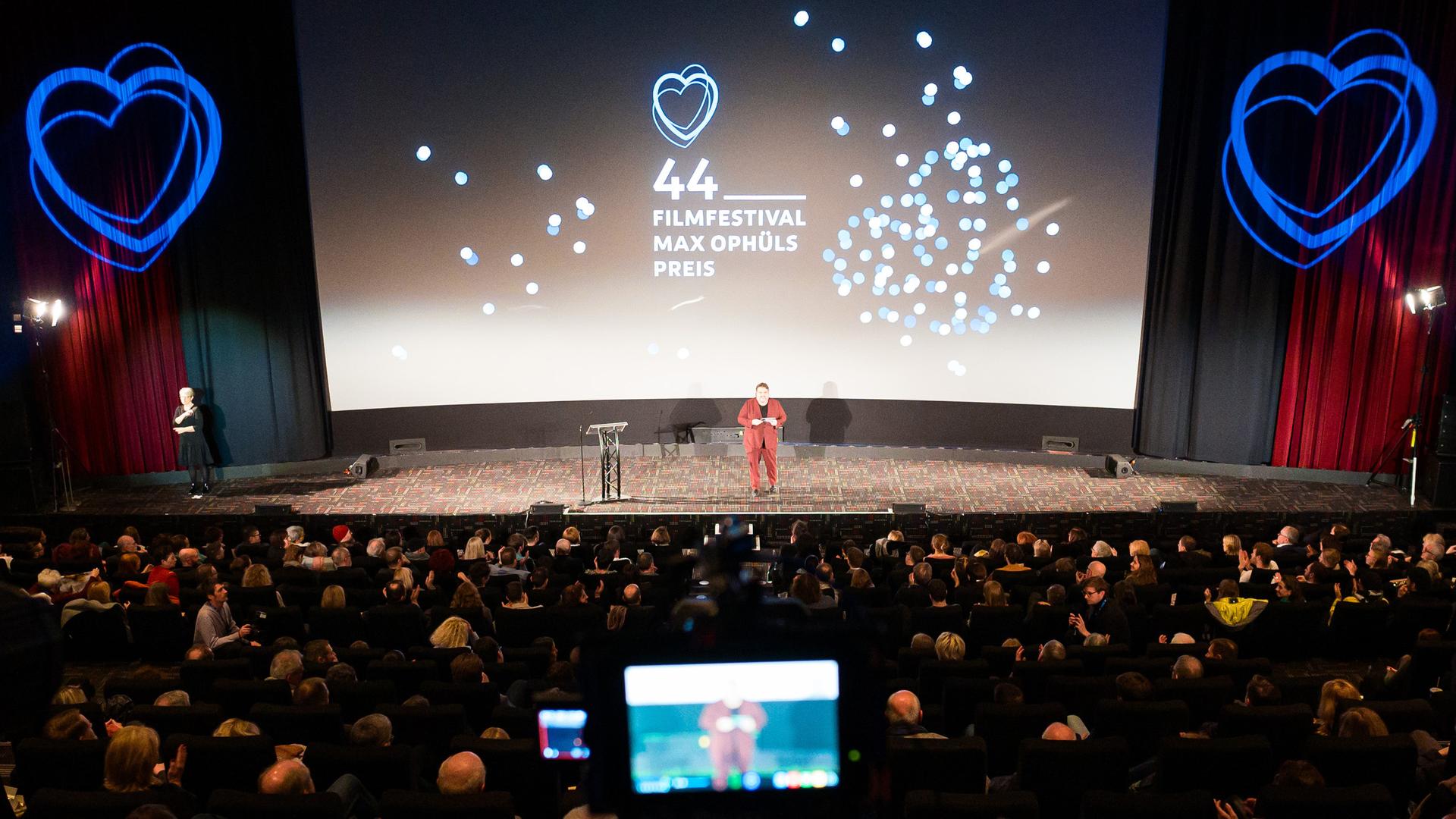 Die Bühne bei der Eröffnung des 44. Filmfestival Max Ophüls Preis, umgeben von Publikum. Im Hintergrund eine Projektion mit Herzen und dem Schrfitzug "44. Filmfestival Max Ophüls Preis"