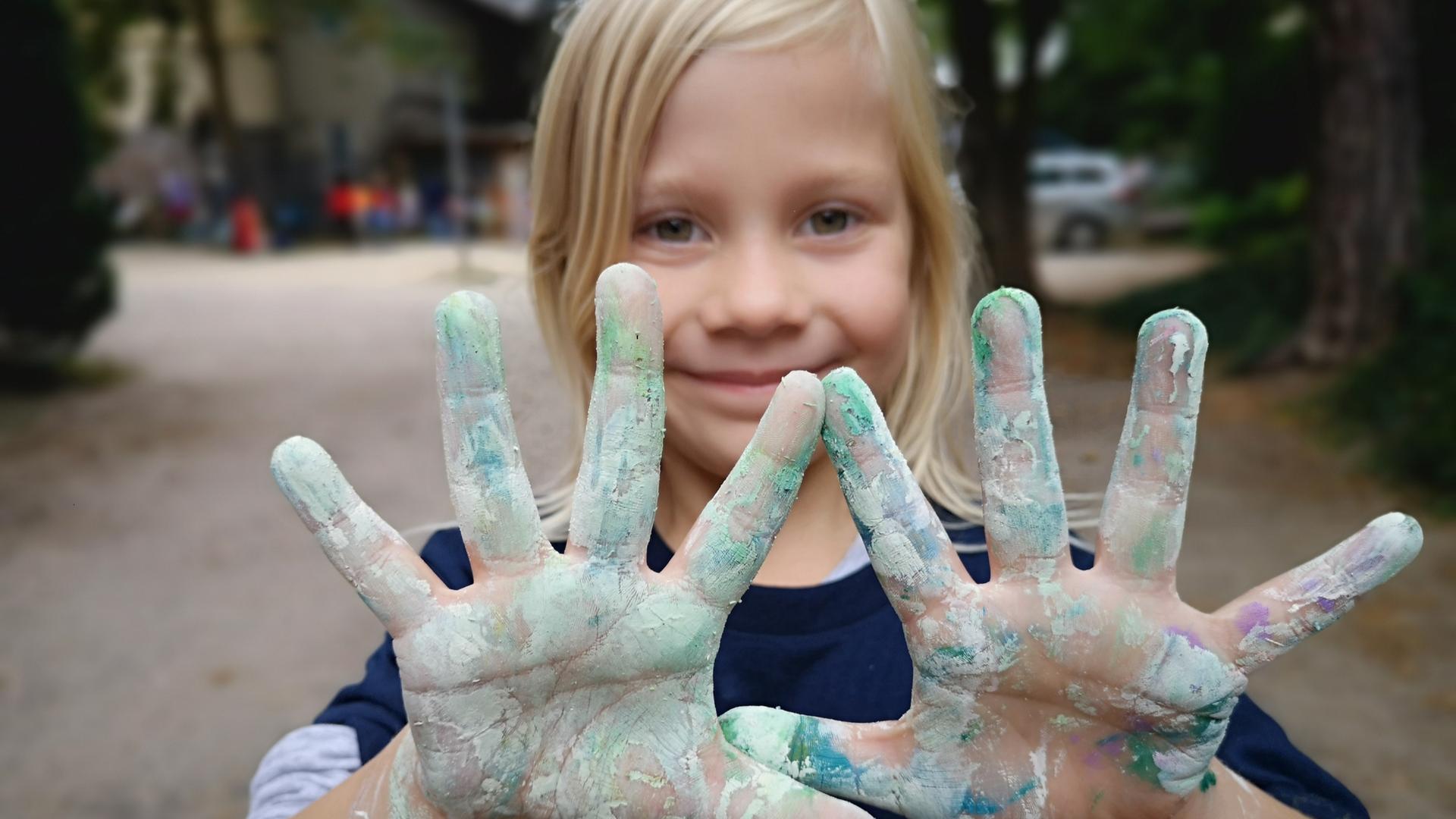 Ein kleines blondes Mädchen hält dem Betrachter ihre mit heller Farbe beschmierten Handflächen entgegen. Sie lächelt. Es ist die Tochter der Fotografin.