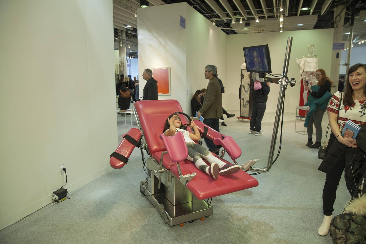 Ein pinker Untersuchungsstuhl steht in einem Ausstellungsraum mit Besucherinnen und Besuchern: Eine Arbeit von Tabita Rezaire bei einer Ausstellung in New York 2018. Ein pinker Untersuchungsstuhl steht in einem Ausstellungsraum mit Besucherinnen und Besuchern: Eine Arbeit von Tabita Rezaire bei einer Ausstellung in New York 2018.