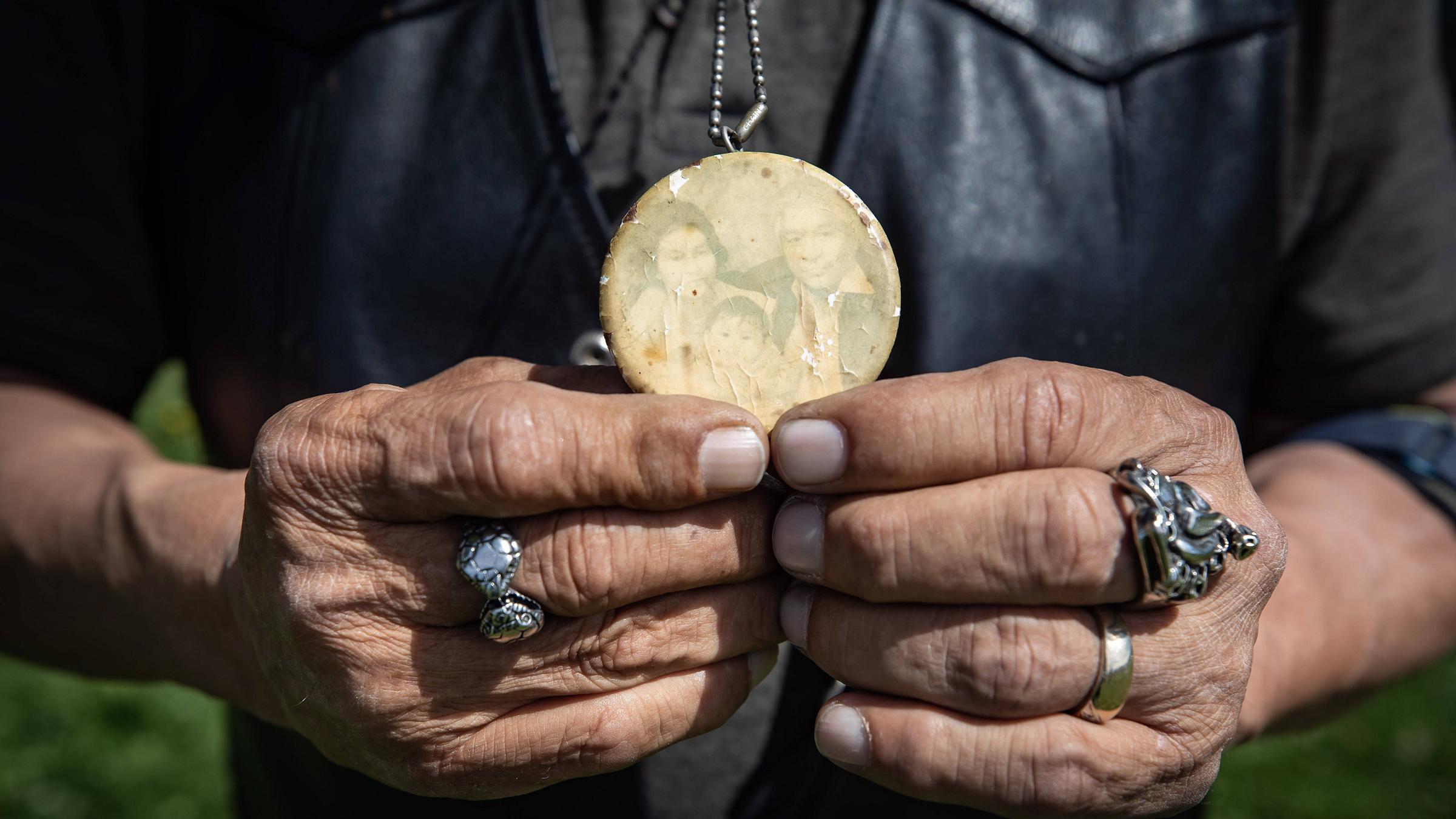 Alte faltige Hände halten ein verblasstes Foto-Medaillon. Darauf ist ein indigenes Paar und ein kleines Mädchen zu sehen. 
