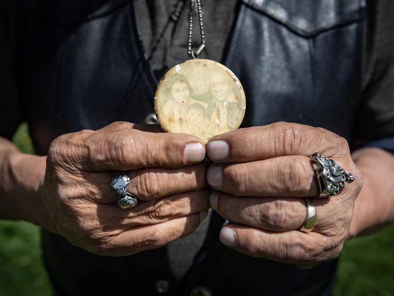 Alte faltige Hände halten ein verblasstes Foto-Medaillon. Darauf ist ein indigenes Paar und ein kleines Mädchen zu sehen. 
