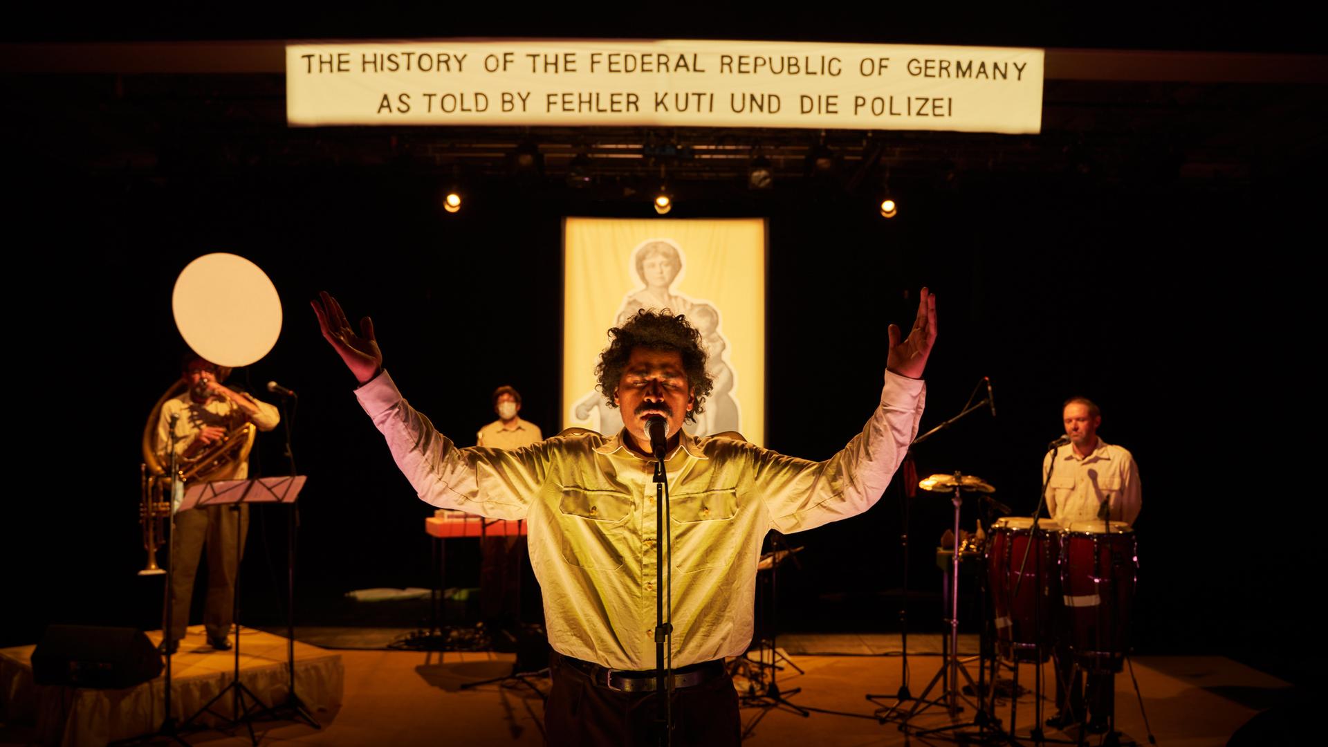 Ein Mann steht mit ausgebreiteten Armen am Mikrofon, hinter ihm steht auf einem Banner: "The History of the Federal Republic of Germany As Told By Fehler Kuti und die Polizei". Ein Mann steht mit ausgebreiteten Armen am Mikrofon, hinter ihm steht auf einem Banner: "The History of the Federal Republic of Germany As Told By Fehler Kuti und die Polizei".