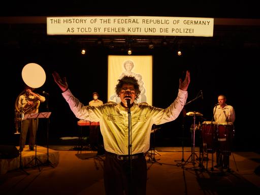 Ein Mann steht mit ausgebreiteten Armen am Mikrofon, hinter ihm steht auf einem Banner: "The History of the Federal Republic of Germany As Told By Fehler Kuti und die Polizei".
