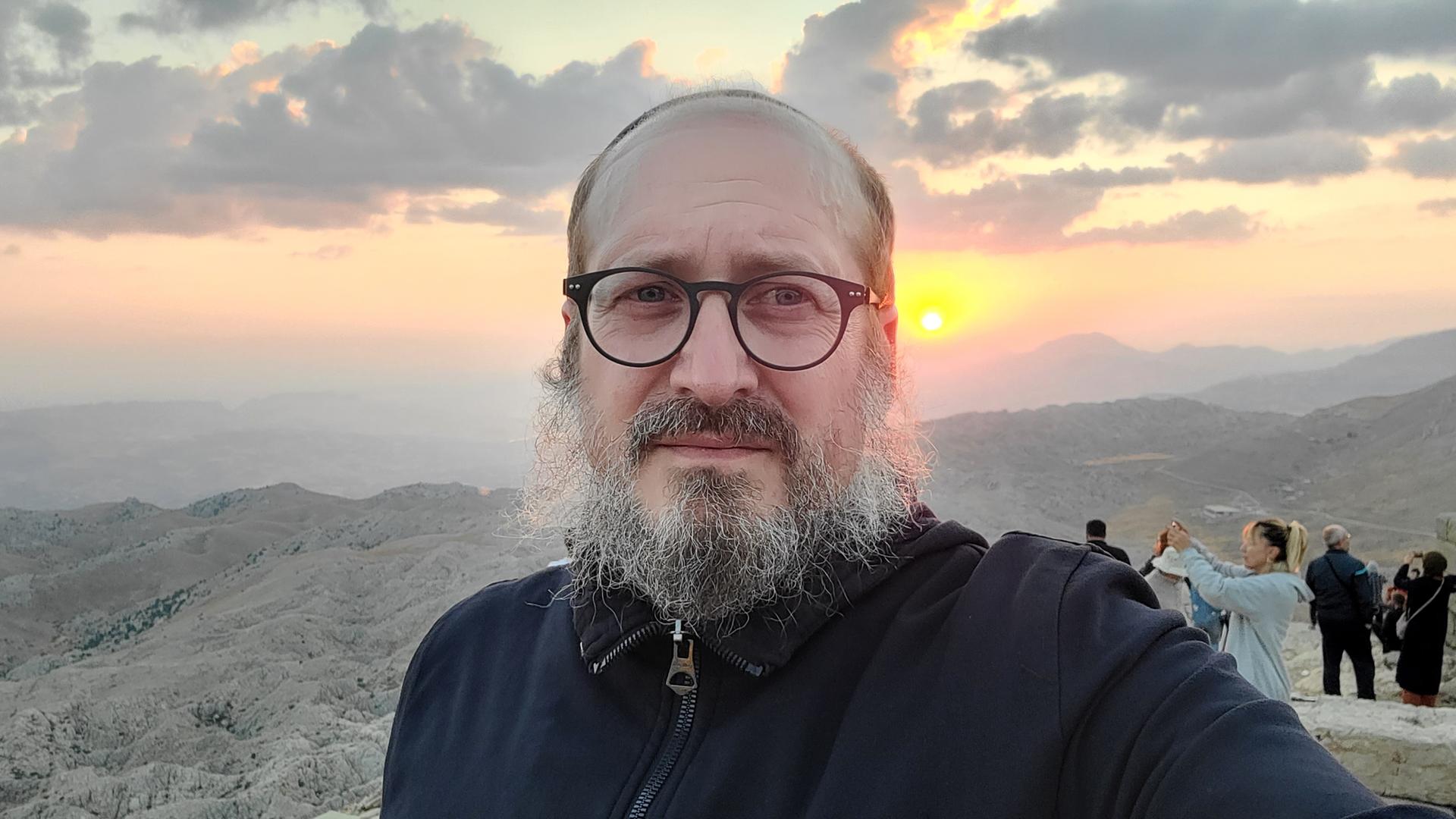 Ein Mann um die 50, Brille, grau melierter Bart, mit Kippa, in einem schwarzen Kapuzenpulli, fotografiert sich selbst – im Hintergrund eine Berglandschaft im Sonnenaufgang und weitere Reisende. Ein Mann um die 50, Brille, grau melierter Bart, mit Kippa, in einem schwarzen Kapuzenpulli, fotografiert sich selbst – im Hintergrund eine Berglandschaft im Sonnenaufgang und weitere Reisende.