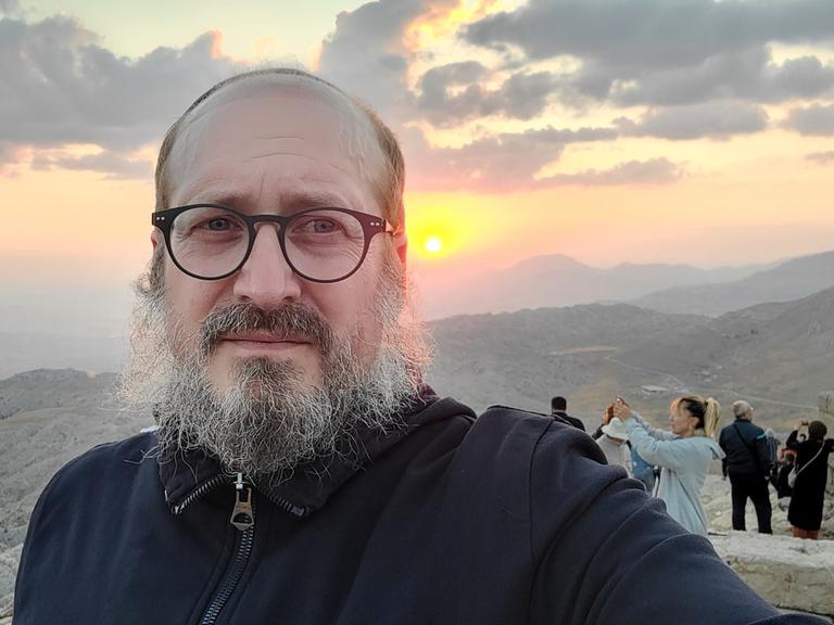 Ein Mann um die 50, Brille, grau melierter Bart, mit Kippa, in einem schwarzen Kapuzenpulli, fotografiert sich selbst – im Hintergrund eine Berglandschaft im Sonnenaufgang und weitere Reisende.