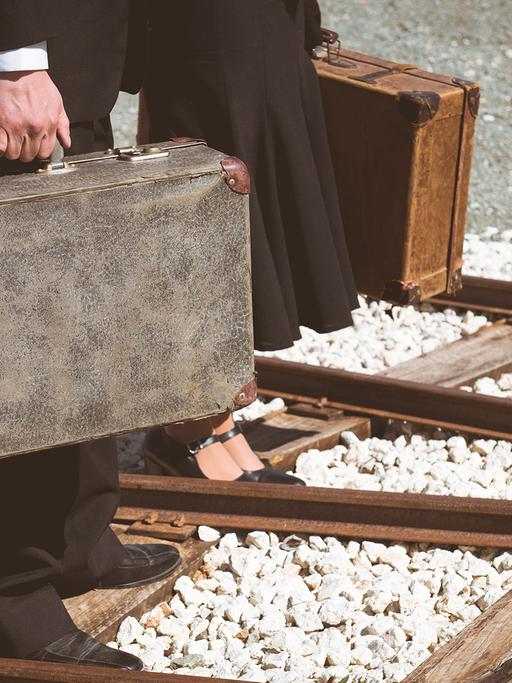 Ein Mann und eine Frau mit je einem Koffer in der Hand, laufen über Bahngleisen. 