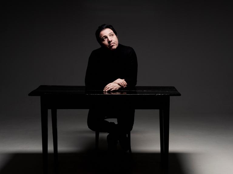 Ein Mann sitzt in einem kahlen Raum an einem Tisch und blickt verträumt an die Decke. 