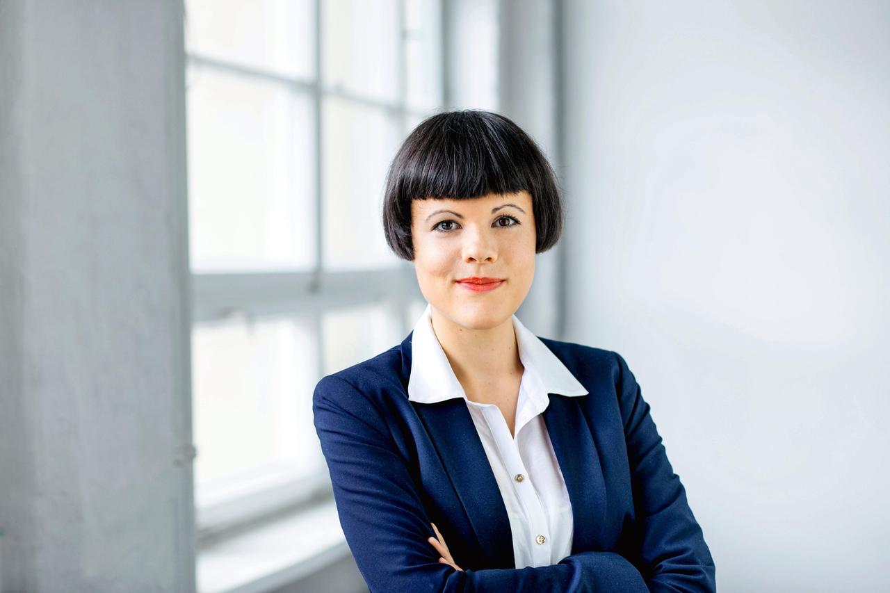 Dana Buchzik im Porträt. Eine junge Frau in einem blauen Blazer steht vor einem Fenster und blickt in die Kamera. Dana Buchzik im Porträt. Eine junge Frau in einem blauen Blazer steht vor einem Fenster und blickt in die Kamera.