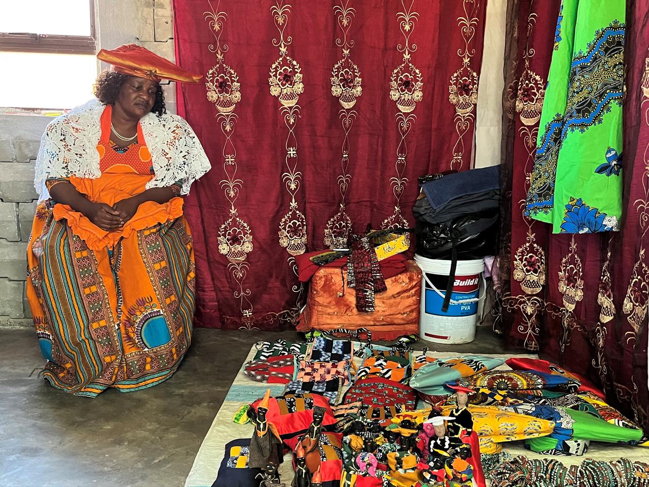 Claudia Kavita in Herero-Tracht: ein ausladendes orangefarbenes Kleid mit Schürze und die typische Kopfbedeckung, die Rinderhörner und damit den Stolz der traditionellen Viehzüchter symbolisiert. An der Wand hängen handgefertigte Kleider, auf dem Boden liegen Kissen, Taschen und Puppen ebenfalls in Herero-Tracht. Claudia Kavita in Herero-Tracht: ein ausladendes orangefarbenes Kleid mit Schürze und die typische Kopfbedeckung, die Rinderhörner und damit den Stolz der traditionellen Viehzüchter symbolisiert. An der Wand hängen handgefertigte Kleider, auf dem Boden liegen Kissen, Taschen und Puppen ebenfalls in Herero-Tracht.