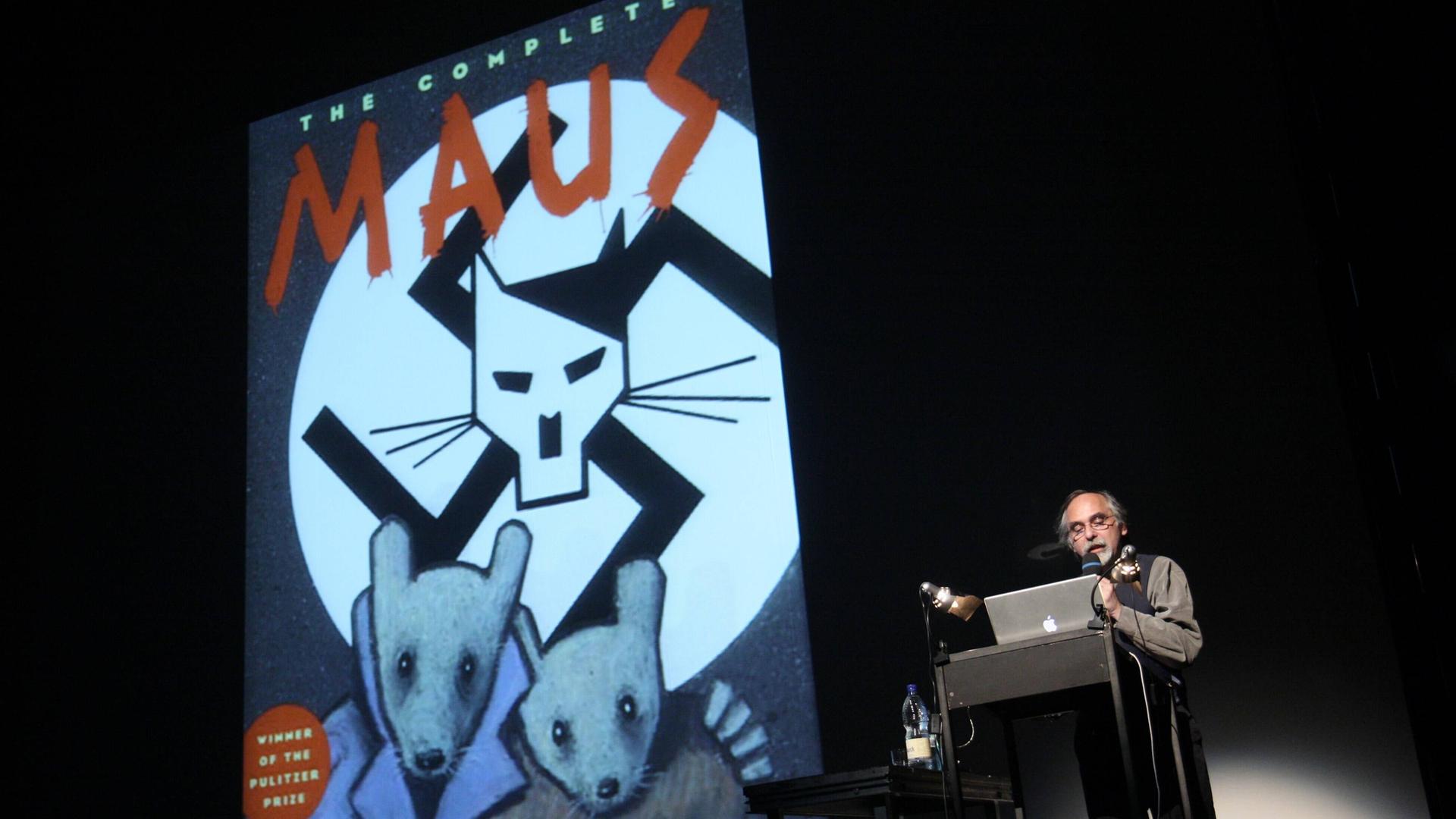 Ein an die Wand projiziertes Comiccover. Oben steht "The complete Maus". Darunter ein gemalter Katzenkopf mit Hitlerbart vor einem Hakenkreuz. Darunter zwei Comicmäuse. Daneben steht ein Mann am Pult, schaut in seinen Computer und spricht in ein Mikrofon. Ein an die Wand projiziertes Comiccover. Oben steht "The complete Maus". Darunter ein gemalter Katzenkopf mit Hitlerbart vor einem Hakenkreuz. Darunter zwei Comicmäuse. Daneben steht ein Mann am Pult, schaut in seinen Computer und spricht in ein Mikrofon.