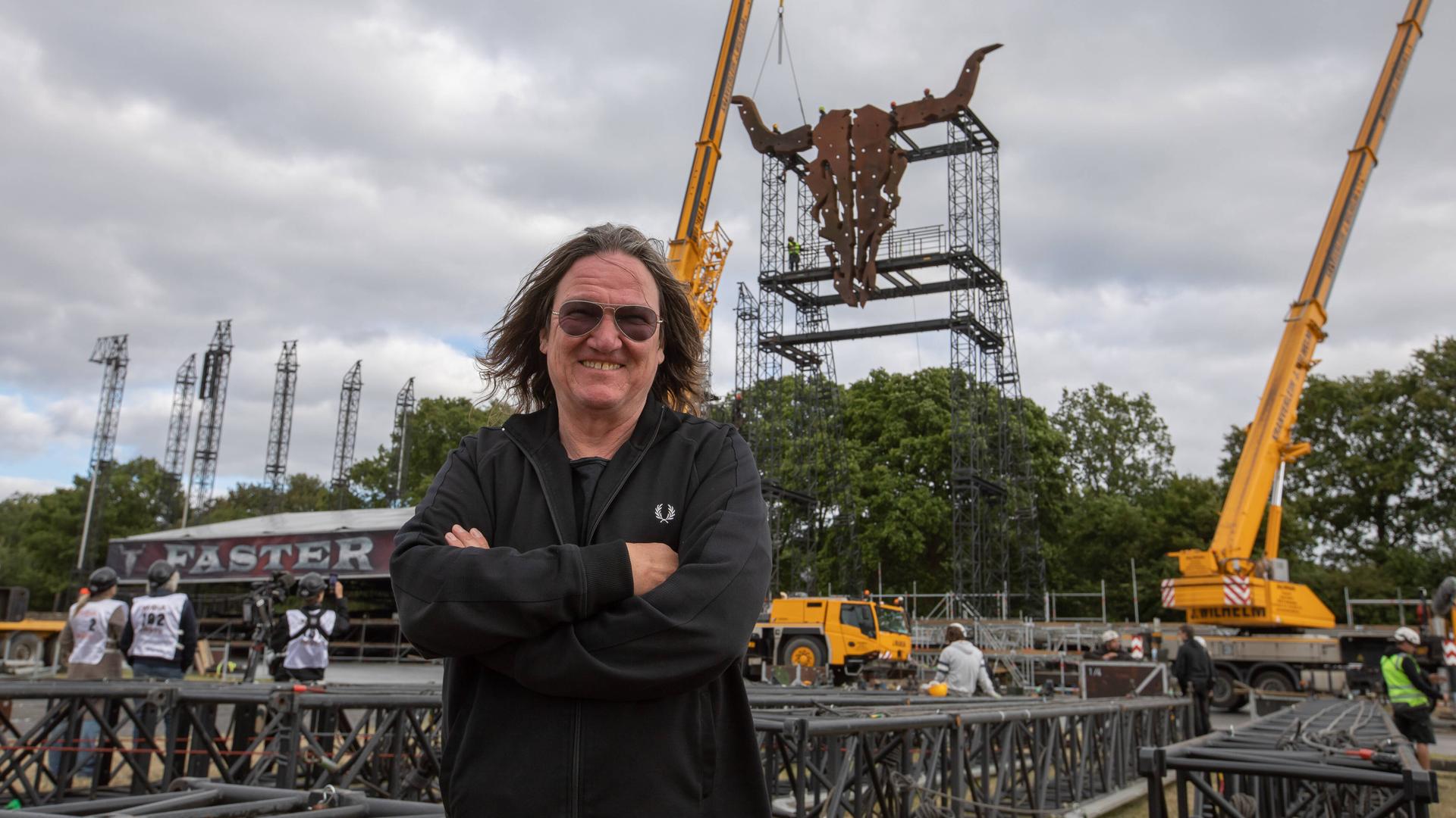 Thomas Jensen steht vor den aufwändigen Aufbauarbeiten des Festivalgeländes. Hinter ihm ist ein großer, aus rostigem Metall gestalteter Kuhschädel zu sehen, der an einer turmhohen Metallkonstruktion aufgehängt ist. Thomas Jensen steht vor den aufwändigen Aufbauarbeiten des Festivalgeländes. Hinter ihm ist ein großer, aus rostigem Metall gestalteter Kuhschädel zu sehen, der an einer turmhohen Metallkonstruktion aufgehängt ist.