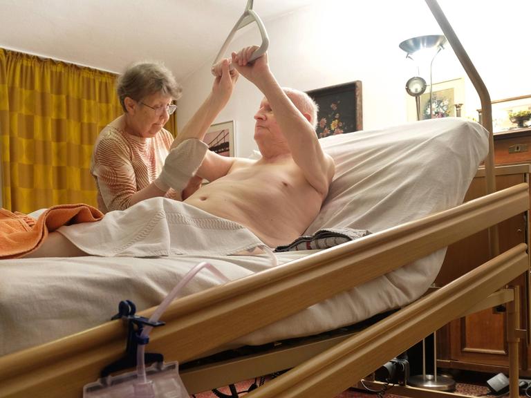 Eine Frau um die 80 wäscht in der gemeinsamen Wohnung ihren Ehemann, der sich im Pflegebett mit Hilfe eines über ihm hängenden Griffs aufrichtet.