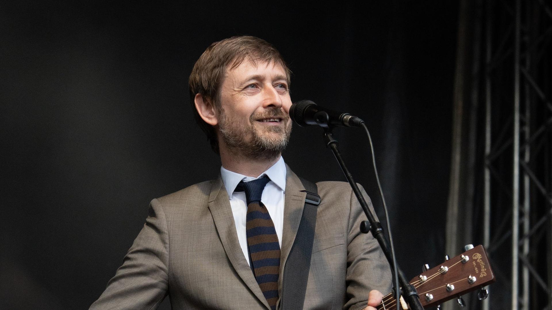 Ein Mann in einem grauen Anzug steht vor einem Mikrofon, er hält eine Gitarre in den Händen.