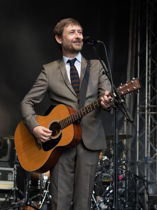 Ein Mann in einem grauen Anzug steht vor einem Mikrofon, er hält eine Gitarre in den Händen.