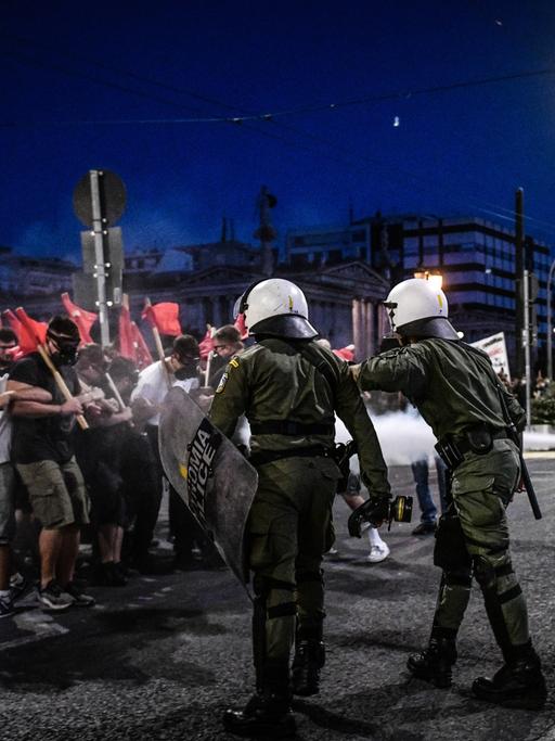 Demonstrierende mit roten Fahnen und Polizisten mit Helmen und Schutzschilden treffen auf einer nächtlichen Straße aufeinander.