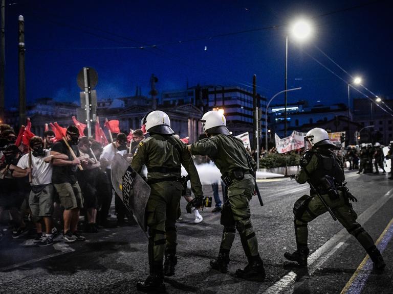 Demonstrierende mit roten Fahnen und Polizisten mit Helmen und Schutzschilden treffen auf einer nächtlichen Straße aufeinander.