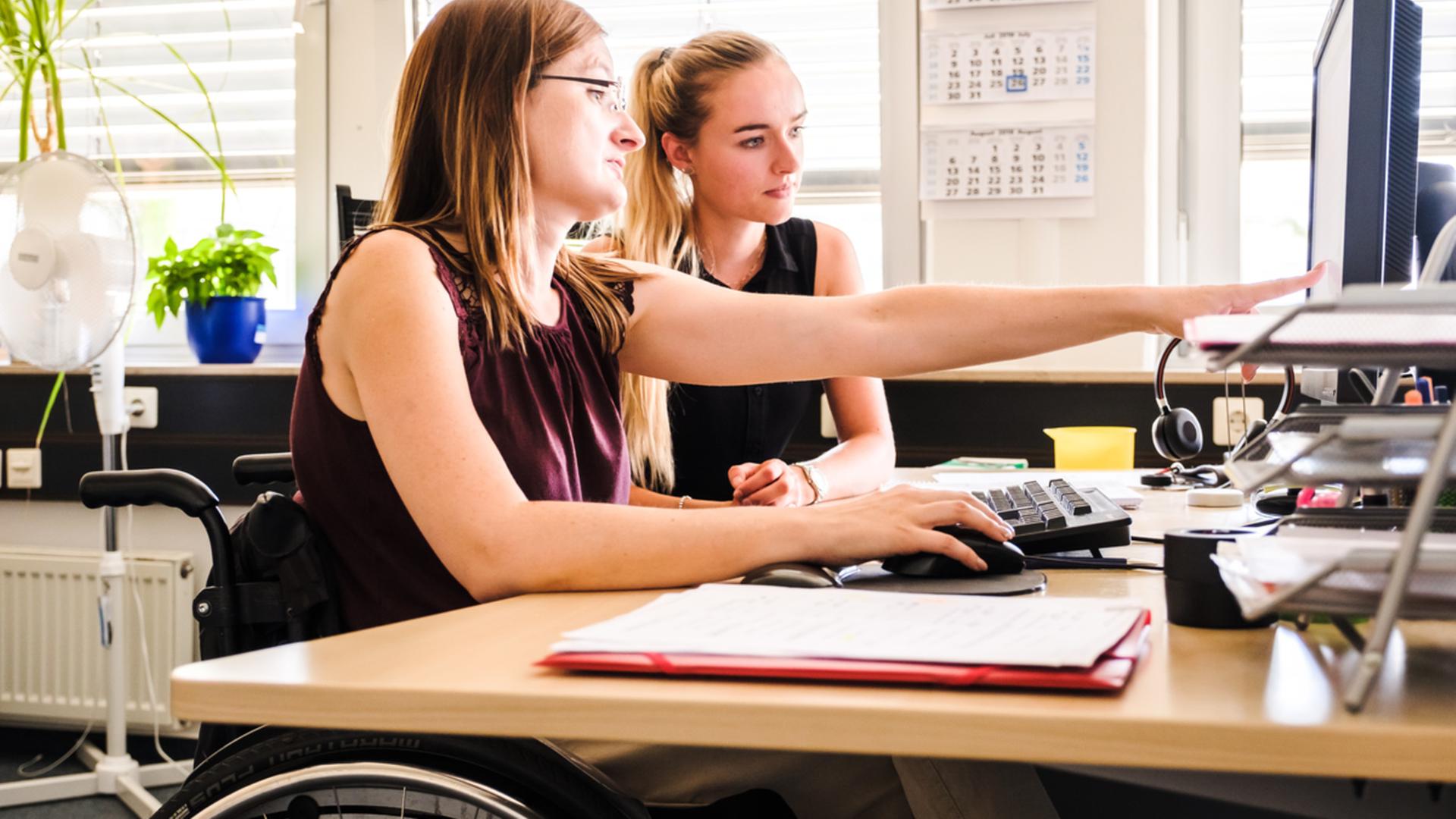 Eine Frau im Rollstuhl zeigt einer anderen Frau etwas am Computer. Eine Frau im Rollstuhl zeigt einer anderen Frau etwas am Computer.