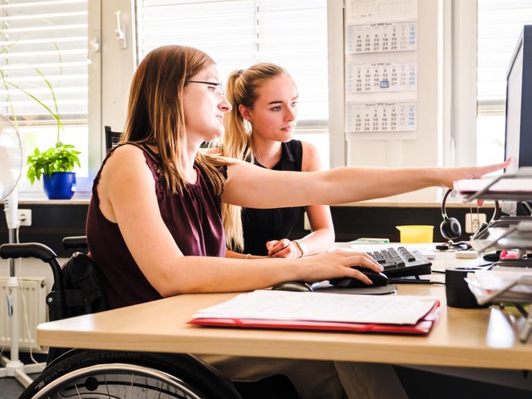 Eine Frau im Rollstuhl zeigt einer anderen Frau etwas am Computer.
