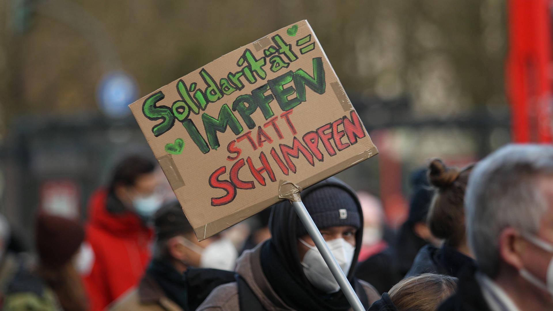 Auf einer Demonstration wird ein Schild präsentiert, auf dem steht: "Solidarität = Impfen statt Schimpfen". Auf einer Demonstration wird ein Schild präsentiert, auf dem steht: "Solidarität = Impfen statt Schimpfen".