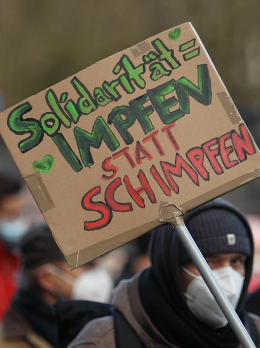 Bei einer Demonstration hält ein Mann ein Schild in die Luft, auf dem steht: "Solidarität = Impfen statt Schimpfen". 