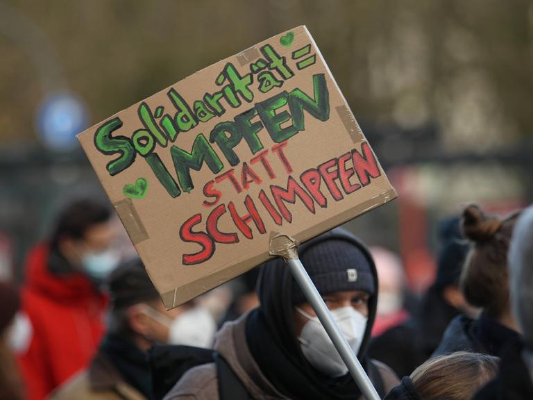 Auf einer Demonstration wird ein Schild präsentiert, auf dem steht: "Solidarität = Impfen statt Schimpfen". 