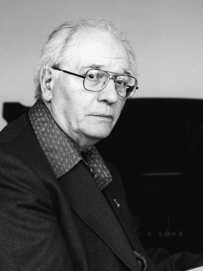 Olivier Messiaen blickt ernst durch seine Brille direkt in die Kamera, während er vor einem Flügel sitzt.