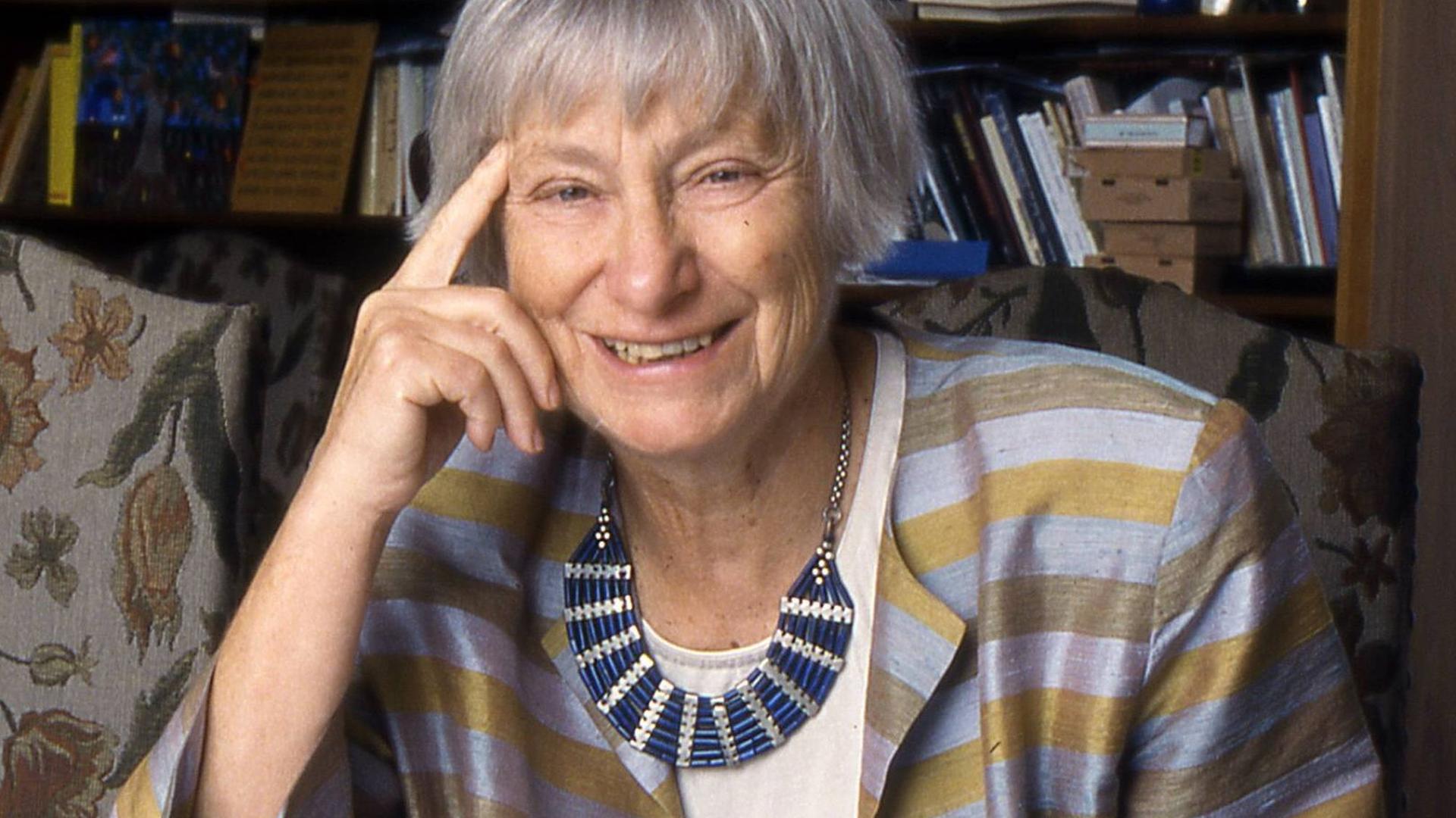 Dorothee Sölle, eine ältere Frau in einer blau-golden gestreiften Jacke, sitzt lächelnd an ihrem Schreibtisch, vor sich verschiedene Schriftstücke ausgebreitet.