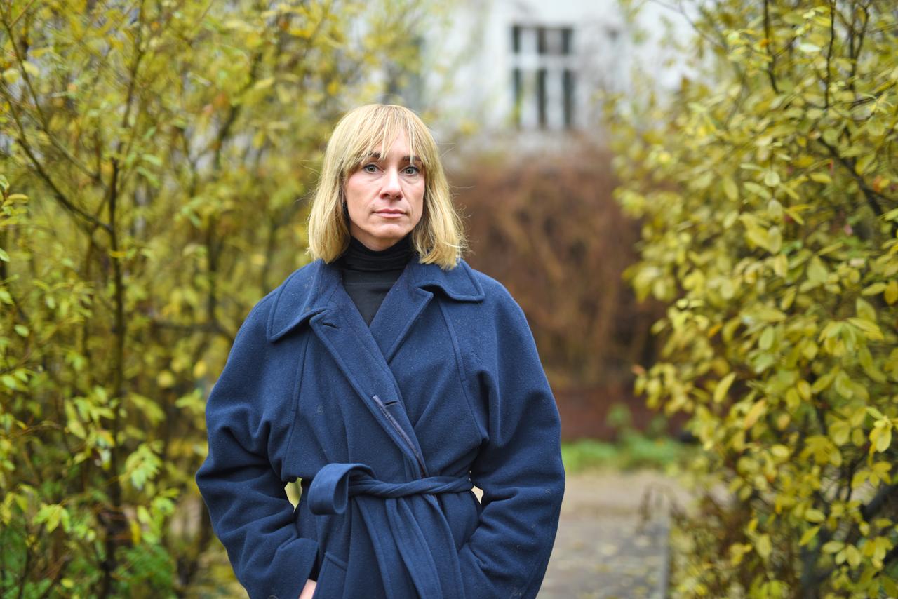 Porträtfoto von Zaraz Gutsch, Mitlied der Gruppe Mathilda, einem Zusammenschluss ehemaliger Muehl-Kommune-BewohnerInnen. Zu sehen ist eine Frau mit blonden Haaren und blauem Mantel zwischen zwei Sträuchern.